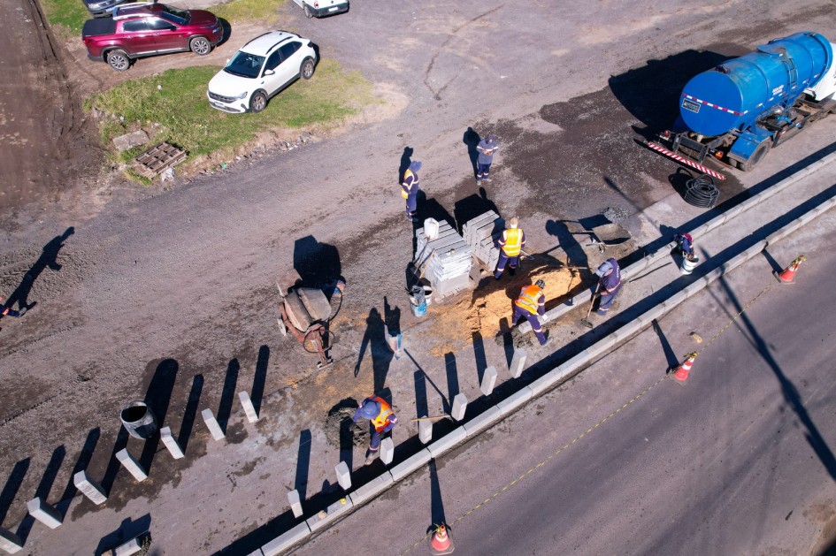 Vista de cima registra trabalhadores em atividade sobre a pista. Eles manuseiam blocos de concreto, pás e carrinhos de obra para instalar meios-fios. Um caminhão azul-petróleo de apoio está estacionado ao lado, enquanto cones delimitam a área de trabalho no asfalto.