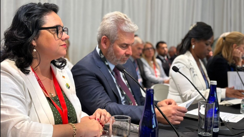 A imagem mostra a secretária do Meio Ambiente, Marjorie Kauffmann, em primeiro plano, sentada à mesa durante reunião ministerial na Semana do Clima de Nova Iorque. Ela veste blazer branco e crachá vermelho, com semblante atento, acompanhada por outros representantes internacionais, incluindo homens e mulheres em trajes formais. A mesa está equipada com microfones, copos d’água e garrafas azuis. Ao fundo, outros participantes aparecem desfocados.