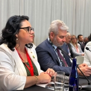 A imagem mostra a secretária do Meio Ambiente, Marjorie Kauffmann, em primeiro plano, sentada à mesa durante reunião ministerial na Semana do Clima de Nova Iorque. Ela veste blazer branco e crachá vermelho, com semblante atento, acompanhada por outros representantes internacionais, incluindo homens e mulheres em trajes formais. A mesa está equipada com microfones, copos d’água e garrafas azuis. Ao fundo, outros participantes aparecem desfocados.