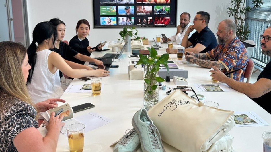 Reunião em andamento em uma sala de escritório moderna. Oito pessoas estão sentadas ao redor de uma mesa branca retangular, com notebooks, celulares, papéis e copos com bebida à frente. No centro da mesa há plantas em vasos transparentes. Ao fundo, uma grande tela exibe uma interface de streaming com miniaturas de filmes e séries. No primeiro plano, em cima da mesa, há um tênis esportivo claro e uma sacola bege com a frase “Music Inspires the Future”. O ambiente é iluminado por luz natural vinda de janelas amplas à direita.