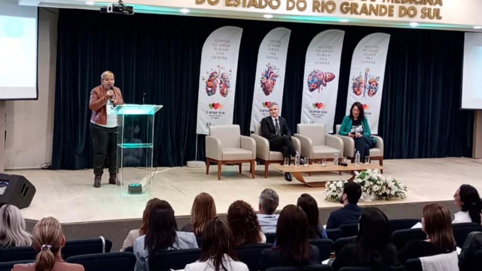 Em um auditório lotado, uma mulher está de pé no palco, falando ao microfone. Há quatro poltronas ao lado, com duas ocupadas por um homem e uma mulher sentados. Ao fundo, há banners que remetem à doação de órgãos, com a marca do governo do Estado. 