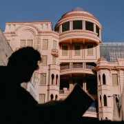 Na imagem, a fachada rosa do prédio histórico da Casa de Cultura Mario Quintana (antigo Hotel Majestic) ocupa o fundo sob céu azul. Em primeiro plano, aparece a silhueta desfocada de uma pessoa segurando um livro aberto, criando contraste entre sombra e luz.