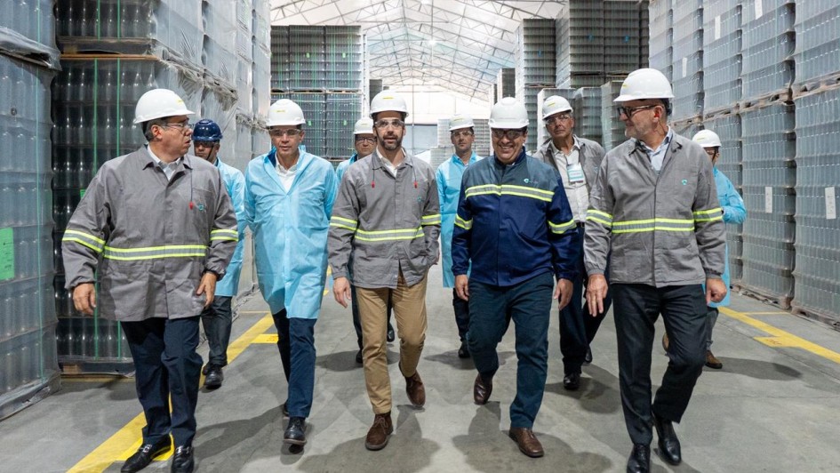 Eduardo Leite aparece no centro, com capacete branco e uniforme cinza, acompanhado por executivos em visita técnica à fábrica.