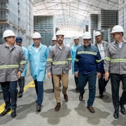 Eduardo Leite aparece no centro, com capacete branco e uniforme cinza, acompanhado por executivos em visita técnica à fábrica.