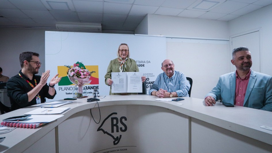 A imagem mostra uma cerimônia oficial em uma sala de reuniões da Secretaria da Saúde do Rio Grande do Sul. Quatro pessoas estão presentes: um homem de óculos e camisa preta à esquerda aplaudindo, Arita exibindo um documento, um homem idoso de camisa xadrez azul e branca sentado, e outro homem de blazer azul claro à direita. Ao fundo há uma apresentação projetada com o logo do "Plano Rio Grande" e texto sobre a Secretaria da Saúde. A mesa curva tem o logo "RS" da Secretaria da Saúde. O ambiente tem teto rebaixado com placas brancas e iluminação fluorescente.