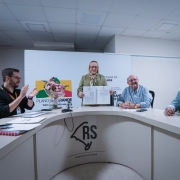 A imagem mostra uma cerimônia oficial em uma sala de reuniões da Secretaria da Saúde do Rio Grande do Sul. Quatro pessoas estão presentes: um homem de óculos e camisa preta à esquerda aplaudindo, Arita exibindo um documento, um homem idoso de camisa xadrez azul e branca sentado, e outro homem de blazer azul claro à direita. Ao fundo há uma apresentação projetada com o logo do "Plano Rio Grande" e texto sobre a Secretaria da Saúde. A mesa curva tem o logo "RS" da Secretaria da Saúde. O ambiente tem teto rebaixado com placas brancas e iluminação fluorescente.