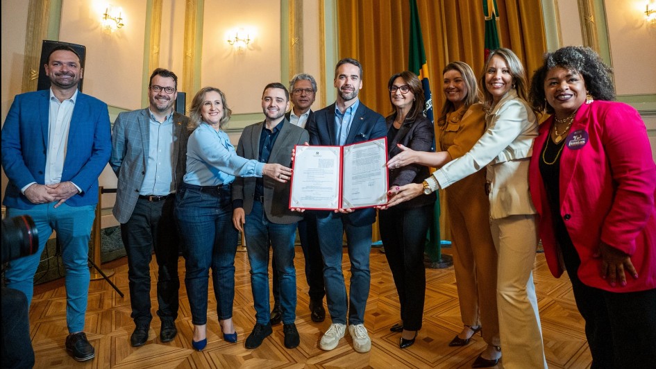 O governador Eduardo Leite e a secretária da Mulher, enfermeira Fábia Richter, estão ao centro da imagem segurando juntos um documento aberto, acompanhados de outras autoridades e representantes. O grupo, composto por homens e mulheres, forma uma linha em pé no salão, diante de cortinas douradas e bandeiras oficiais do Brasil e do Rio Grande do Sul.