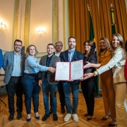 O governador Eduardo Leite e a secretária da Mulher, enfermeira Fábia Richter, estão ao centro da imagem segurando juntos um documento aberto, acompanhados de outras autoridades e representantes. O grupo, composto por homens e mulheres, forma uma linha em pé no salão, diante de cortinas douradas e bandeiras oficiais do Brasil e do Rio Grande do Sul.