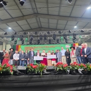 Na imagem, um grupo de pessoas posa no palco durante cerimônia de premiação da campanha Ganha-Ganha Farroupilha. Homens e mulheres, alguns em trajes típicos gaúchos, estão alinhados em frente ao painel com a identidade visual da campanha. Entre eles, participantes exibem certificado e cheque simbólico, enquanto o cenário inclui iluminação de palco e arranjos florais vermelhos à frente.