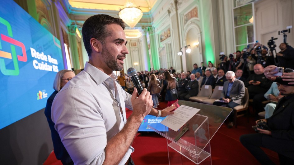 Evento formal em salão elegante. Homem fala ao microfone num púlpito transparente, com tela atrás exibindo “Rede Bem Cuidar RS”. Público atento, câmeras registrando. Parece anúncio oficial sobre saúde no Rio Grande do Sul.