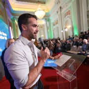 Evento formal em salão elegante. Homem fala ao microfone num púlpito transparente, com tela atrás exibindo “Rede Bem Cuidar RS”. Público atento, câmeras registrando. Parece anúncio oficial sobre saúde no Rio Grande do Sul.