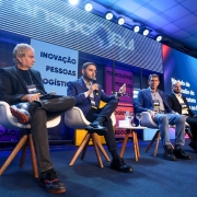 Na imagem, quatro homens estão sentados em cadeiras brancas sobre um palco, participando de um painel da TranspoSul. O vice-governador do Rio Grande do Sul, Gabriel Souza, aparece em destaque ao centro, vestindo terno azul e segurando um microfone enquanto fala ao público. Atrás, há painéis coloridos com palavras como "Inovação", "Pessoas" e "Logística". O ambiente é iluminado com luzes de palco, e uma tela grande exibe informações sobre o evento.
