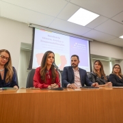 Cinco mulheres e um homem compõem uma mesa de debates. Uma das mulheres fala ao microfone, enquanto os demais escutam. Ao fundo, há um painel com a marca do Seminário Gênero, Justiça e Responsabilização. 