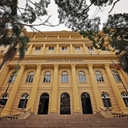 A imagem retrata a fachada imponente de um edifício histórico identificado como “SECRETARIA DA FAZENDA”. A construção apresenta elementos clássicos da arquitetura, como colunas altas, janelas em arco e detalhes ornamentais, com tonalidade amarelada.