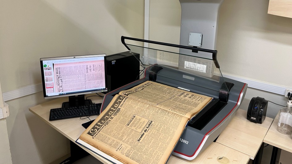 Esta é uma fotografia que mostra um equipamento de digitalização de documentos e um computador, em um ambiente de arquivo ou biblioteca.

No centro da imagem, sobre uma mesa de trabalho clara, está um scanner planetário profissional de cor cinza e bordas vermelhas, da marca ZEUTSCHEL. O scanner possui uma haste superior curva com a marca e uma base plana onde um jornal antigo e amarelado está aberto para digitalização. O título "Literatura" é visível na parte superior do jornal.

À esquerda, sobre a mesa, há um monitor de computador que exibe o resultado da digitalização do jornal, mostrando o texto com colunas escuras. O monitor está conectado a uma unidade de sistema (CPU) preta e um teclado.

A sala tem paredes de cor bege e boa iluminação. No canto inferior direito, há um dispensador de álcool em gel e outros itens. O ambiente é de trabalho e conservação de documentos.