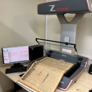 Esta é uma fotografia que mostra um equipamento de digitalização de documentos e um computador, em um ambiente de arquivo ou biblioteca.

No centro da imagem, sobre uma mesa de trabalho clara, está um scanner planetário profissional de cor cinza e bordas vermelhas, da marca ZEUTSCHEL. O scanner possui uma haste superior curva com a marca e uma base plana onde um jornal antigo e amarelado está aberto para digitalização. O título "Literatura" é visível na parte superior do jornal.

À esquerda, sobre a mesa, há um monitor de computador que exibe o resultado da digitalização do jornal, mostrando o texto com colunas escuras. O monitor está conectado a uma unidade de sistema (CPU) preta e um teclado.

A sala tem paredes de cor bege e boa iluminação. No canto inferior direito, há um dispensador de álcool em gel e outros itens. O ambiente é de trabalho e conservação de documentos.