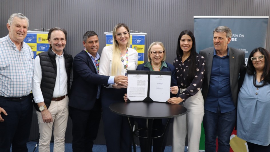 Esta é uma fotografia de oito pessoas (cinco homens e três mulheres) posando atrás de uma pequena mesa redonda preta, em um evento de assinatura de convênio, em Eldorado do Sul.

No centro, a prefeita do município, em camisa branca de mangas longas, e a Secretária de Saúde do Estado, Arita Bergmann, em blazer azul-marinho, seguram um documento assinado sobre a mesa. Elas olham para a câmera e sorriem.

O grupo, composto por autoridades e secretários, está posicionado atrás e ao redor da mesa. Todos sorriem e vestem trajes sociais ou esporte fino.

O fundo é uma parede de painéis cinzas escuros, com dois banners verticais visíveis:

O banner da esquerda, com o logo de Eldorado do Sul, em padrão xadrez azul e amarelo.

O banner da direita, com o logo da Secretaria da Saúde do Governo do Estado do Rio Grande do Sul.