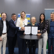 Esta é uma fotografia de oito pessoas (cinco homens e três mulheres) posando atrás de uma pequena mesa redonda preta, em um evento de assinatura de convênio, em Eldorado do Sul.

No centro, a prefeita do município, em camisa branca de mangas longas, e a Secretária de Saúde do Estado, Arita Bergmann, em blazer azul-marinho, seguram um documento assinado sobre a mesa. Elas olham para a câmera e sorriem.

O grupo, composto por autoridades e secretários, está posicionado atrás e ao redor da mesa. Todos sorriem e vestem trajes sociais ou esporte fino.

O fundo é uma parede de painéis cinzas escuros, com dois banners verticais visíveis:

O banner da esquerda, com o logo de Eldorado do Sul, em padrão xadrez azul e amarelo.

O banner da direita, com o logo da Secretaria da Saúde do Governo do Estado do Rio Grande do Sul.