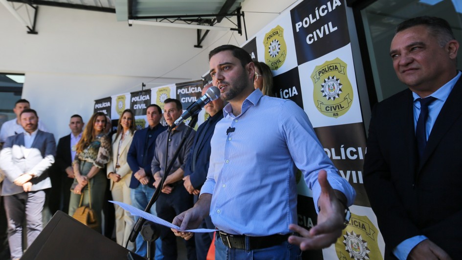 Fotografia de um evento oficial da Polícia Civil do Rio Grande do Sul. Em primeiro plano, um homem — identificado como o vice-governador Gabriel Souza — está falando ao microfone e segurando papéis, aparentemente fazendo um discurso. Ao fundo, há um painel institucional com o logotipo da Polícia Civil e a inscrição “POLÍCIA CIVIL”. Outras pessoas estão posicionadas ao lado e atrás do orador, indicando uma cerimônia formal, possivelmente a inauguração de uma delegacia. O ambiente é interno e bem iluminado, com foco na solenidade e na presença de autoridades.