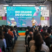 A imagem mostra o governador em um palco durante um evento realizado em um espaço amplo com estilo industrial. Ao fundo, há uma grande tela exibindo os dizeres “RIO GRANDE DO FUTURO” e “5 IDEIAS INEGOCIÁVEIS”. O público está sentado e atento. Banners e sinalizações ao redor do palco incluem o nome “SEMANA CALDEIRA”.