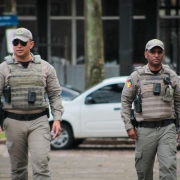 Dois polícias, em plano americano e de frente, caminhando na rua. Eles estão sérios e uniformizados, usando coletes com as câmeras.
