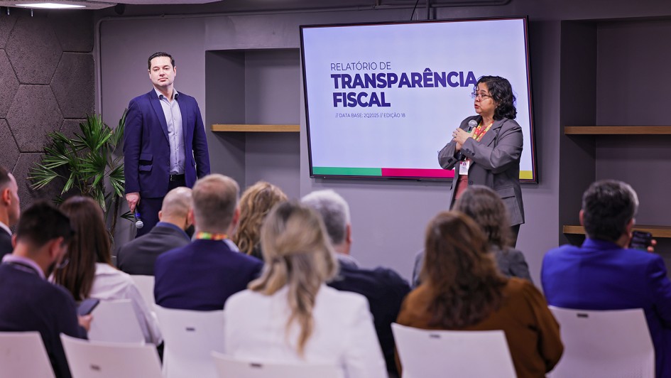 A imagem mostra um ambiente de conferência com várias pessoas sentadas em cadeiras brancas, voltadas para um palco onde dois palestrantes (um homem e uma mulher) estão em ação, ambos utilizando microfone. Ao fundo, uma grande tela exibe o título “RELATÓRIO DE TRANSPARÊNCIA FISCAL”
