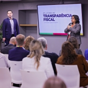 A imagem mostra um ambiente de conferência com várias pessoas sentadas em cadeiras brancas, voltadas para um palco onde dois palestrantes (um homem e uma mulher) estão em ação, ambos utilizando microfone. Ao fundo, uma grande tela exibe o título “RELATÓRIO DE TRANSPARÊNCIA FISCAL”