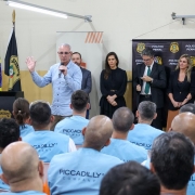 Na imagem, o secretário Jorge Pozzobom fala ao microfone para um grupo de pessoas usando coletes azuis com a inscrição Piccadilly. Atrás dele, ao lado de autoridades e convidados, estão as bandeiras do Rio Grande do Sul, do Brasil e da Polícia Penal. 