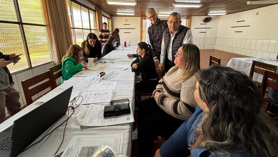 Um grupo de pessoas está reunido em uma sala com teto de madeira e grandes janelas. Elas estão distribuídas ao redor de uma mesa longa, coberta com toalhas brancas, papéis e documentos. Algumas pessoas estão sentadas, outras em pé, envolvidas em discussões ou analisando os materiais sobre a mesa. Há um laptop e outros itens de escritório, como canetas e pastas. O ambiente sugere a realização de uma reunião ou oficina em espaço interno.