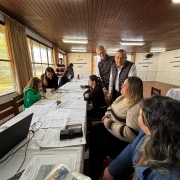 Um grupo de pessoas está reunido em uma sala com teto de madeira e grandes janelas. Elas estão distribuídas ao redor de uma mesa longa, coberta com toalhas brancas, papéis e documentos. Algumas pessoas estão sentadas, outras em pé, envolvidas em discussões ou analisando os materiais sobre a mesa. Há um laptop e outros itens de escritório, como canetas e pastas. O ambiente sugere a realização de uma reunião ou oficina em espaço interno.