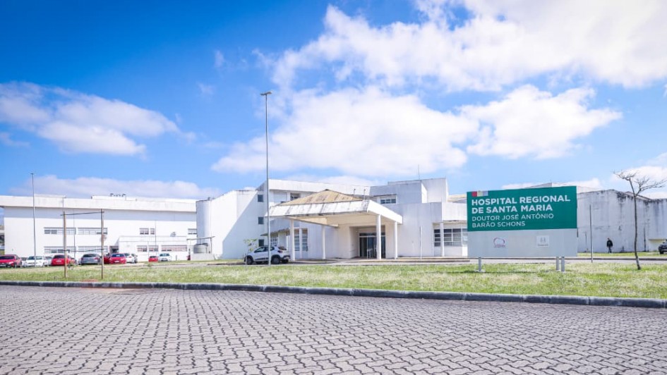 A imagem mostra a fachada do Hospital Regional de Santa Maria, identificado por uma placa verde com letras brancas que diz: “HOSPITAL REGIONAL DE SANTA MARIA”. O prédio é branco, com linhas simples e modernas, e possui uma entrada coberta por uma estrutura em formato triangular. 