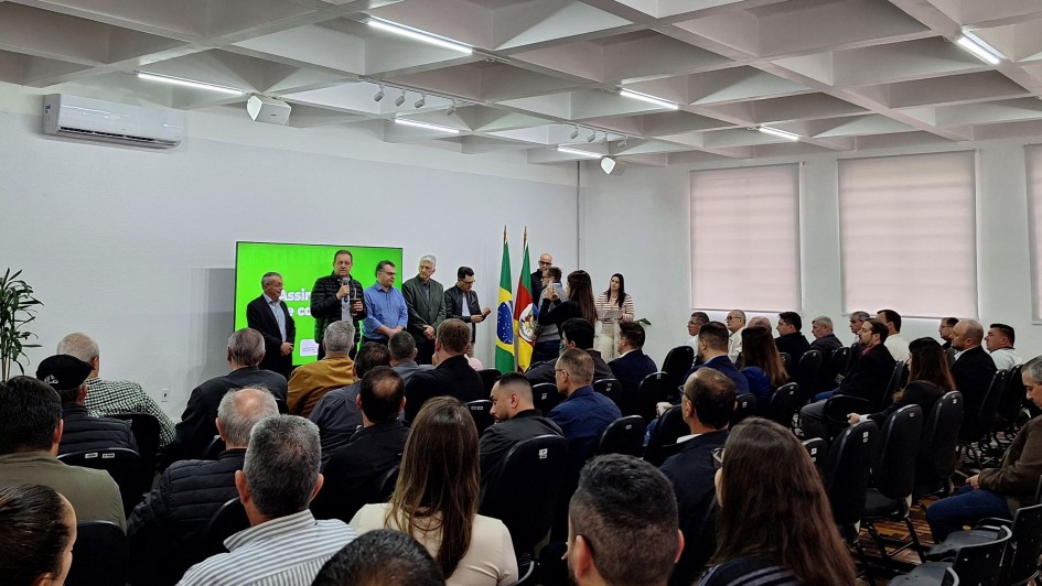 Um grupo de pessoas está reunido em uma sala com paredes brancas e teto com iluminação embutida em painéis quadrados. A sala possui fileiras de cadeiras, a maioria ocupada por pessoas sentadas voltadas para a frente. Na parte frontal do ambiente, há um telão verde com texto projetado e várias pessoas em pé, algumas segurando microfones. Duas bandeiras estão posicionadas próximas aos palestrantes. A cena retrata um evento formal ou uma apresentação pública.