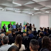 Um grupo de pessoas está reunido em uma sala com paredes brancas e teto com iluminação embutida em painéis quadrados. A sala possui fileiras de cadeiras, a maioria ocupada por pessoas sentadas voltadas para a frente. Na parte frontal do ambiente, há um telão verde com texto projetado e várias pessoas em pé, algumas segurando microfones. Duas bandeiras estão posicionadas próximas aos palestrantes. A cena retrata um evento formal ou uma apresentação pública.