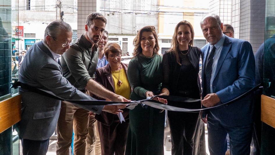 Um grupo de sete pessoas está reunido em uma cerimônia de inauguração, cortando uma fita preta com laço branco. Todos estão sorrindo e posicionados próximos uns dos outros. A maioria está vestindo trajes formais — ternos e vestidos. O local parece ser a entrada de um prédio moderno, com revestimento de pastilhas claras. Ao fundo, pela porta de vidro, é possível ver uma rua movimentada, com prédios antigos, lojas e fiação elétrica. A cerimônia parece contar com a presença de autoridades ou convidados importantes.