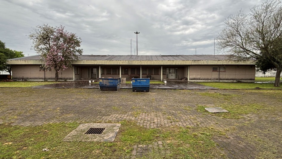 A imagem apresenta a fachada de um prédio térreo, com estrutura alongada e paredes em tom bege. Em frente ao edifício, há um pátio pavimentado, parcialmente coberto por grama, e duas grandes caçambas azuis posicionadas no centro. 
