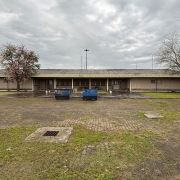 A imagem apresenta a fachada de um prédio térreo, com estrutura alongada e paredes em tom bege. Em frente ao edifício, há um pátio pavimentado, parcialmente coberto por grama, e duas grandes caçambas azuis posicionadas no centro. 
