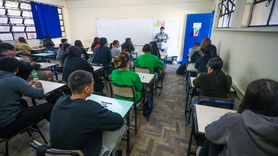Foto mostra sala de aula com alunos acompanhando aula de professora. 
