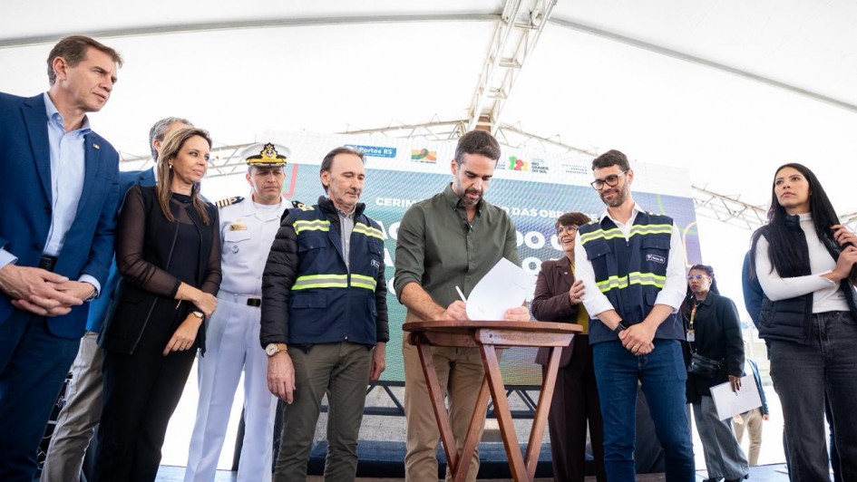 A imagem mostra uma cerimônia oficial realizada sob uma tenda branca, com um grupo de pessoas posicionadas sobre um palco. No centro, há uma mesa de madeira onde o governador assina um documento, enquanto outros observam.