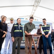 A imagem mostra uma cerimônia oficial realizada sob uma tenda branca, com um grupo de pessoas posicionadas sobre um palco. No centro, há uma mesa de madeira onde o governador assina um documento, enquanto outros observam.