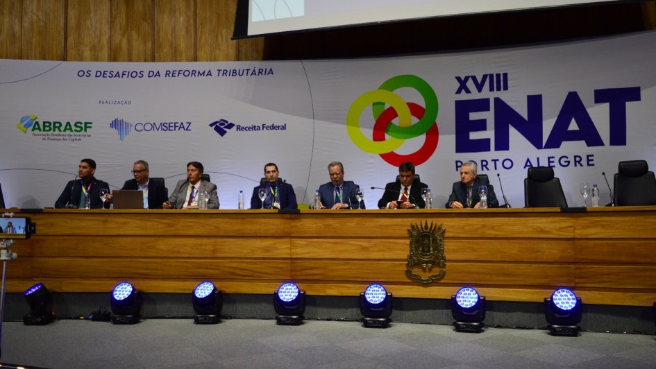 A imagem mostra um auditório com um painel de debates composto por sete pessoas sentadas atrás de uma longa mesa de madeira, sobre um palco. Ao fundo, há um grande painel com o logotipo do evento “XVIII ENAT – Porto Alegre” e o tema “Os desafios da reforma tributária”.