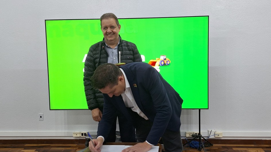 Um homem está assinando um documento sobre uma mesa, enquanto outro está em pé, atrás dele, sorrindo para a foto. Ao fundo, há uma tela verde. 