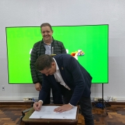 Um homem está assinando um documento sobre uma mesa, enquanto outro está em pé, atrás dele, sorrindo para a foto. Ao fundo, há uma tela verde. 