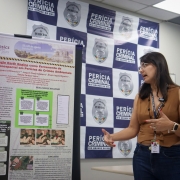 Foto mostra pesquisadora de pé em frente a um banner com trabalho acadêmico fazendo apresentação da pesquisa.