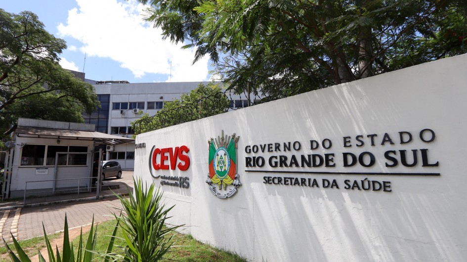 Fachada de prédio governamental com placa em destaque na frente. A placa exibe os textos "CEVS Coordenadoria RS" e "Governo do Estado Rio Grande do Sul Secretaria da Saúde", acompanhados de um brasão colorido. Ao fundo, vê-se um edifício de vários andares com janelas alinhadas horizontalmente. Há árvores e vegetação ao redor, criando um ambiente sombreado e arborizado.
