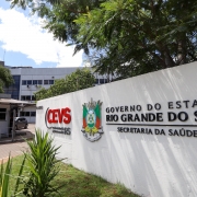 Fachada de prédio governamental com placa em destaque na frente. A placa exibe os textos "CEVS Coordenadoria RS" e "Governo do Estado Rio Grande do Sul Secretaria da Saúde", acompanhados de um brasão colorido. Ao fundo, vê-se um edifício de vários andares com janelas alinhadas horizontalmente. Há árvores e vegetação ao redor, criando um ambiente sombreado e arborizado.