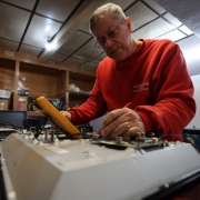 A foto mostra um homem concentrado no conserto de um equipamento eletrônico. Ele usa um multímetro para fazer medições em uma placa de circuito sobre uma bancada de trabalho. Veste moletom vermelho e óculos.