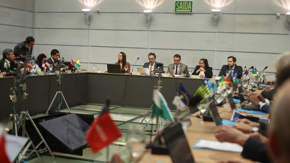 A imagem mostra uma reunião formal em uma sala de conferência. Os participantes, sentados ao redor de uma mesa em formato de “U”, aparentam ser autoridades ou representantes de diferentes estados ou países, o que é sugerido pelas pequenas bandeiras dispostas em frente a cada pessoa. No centro da imagem, há cinco pessoas em destaque, incluindo uma mulher falando ao microfone. Algumas delas estão usando notebooks, papéis e pastas.
Na parte superior da imagem, vê-se uma placa verde com a palavra "SAÍDA", indicando que a reunião ocorre em um local de língua portuguesa — provavelmente no Brasil. Ao fundo e ao lado, há tripés com câmeras ou equipamentos de gravação, sugerindo que o evento está sendo registrado.
O ambiente é moderno, com paredes claras, boa iluminação e uma estrutura de vidro no chão. A presença das bandeiras de diferentes estados brasileiros (como Pernambuco, Ceará, Goiás, entre outras) e de países sul-americanos (como Bolívia e Chile) sugere que esta é uma reunião de caráter interestadual ou internacional, possivelmente relacionada à economia, finanças públicas ou cooperação governamental.
