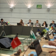 A imagem mostra uma reunião formal em uma sala de conferência. Os participantes, sentados ao redor de uma mesa em formato de “U”, aparentam ser autoridades ou representantes de diferentes estados ou países, o que é sugerido pelas pequenas bandeiras dispostas em frente a cada pessoa. No centro da imagem, há cinco pessoas em destaque, incluindo uma mulher falando ao microfone. Algumas delas estão usando notebooks, papéis e pastas.
Na parte superior da imagem, vê-se uma placa verde com a palavra "SAÍDA", indicando que a reunião ocorre em um local de língua portuguesa — provavelmente no Brasil. Ao fundo e ao lado, há tripés com câmeras ou equipamentos de gravação, sugerindo que o evento está sendo registrado.
O ambiente é moderno, com paredes claras, boa iluminação e uma estrutura de vidro no chão. A presença das bandeiras de diferentes estados brasileiros (como Pernambuco, Ceará, Goiás, entre outras) e de países sul-americanos (como Bolívia e Chile) sugere que esta é uma reunião de caráter interestadual ou internacional, possivelmente relacionada à economia, finanças públicas ou cooperação governamental.