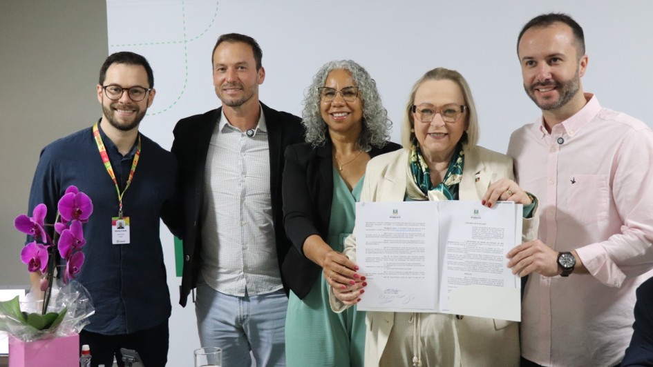 A imagem mostra um grupo de cinco pessoas reunidas em um ambiente interno, posando para uma foto durante um momento formal. No centro, duas pessoas seguram um documento aberto.