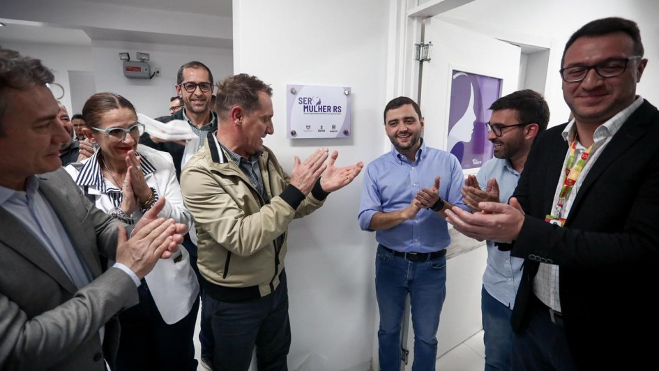 A imagem mostra um grupo de pessoas em um ambiente interno, comemorando a inauguração de um espaço chamado "Ser Mulher RS", conforme indicado pela placa na parede. A placa tem o logotipo do governo do Rio Grande do Sul e de outros órgãos públicos.
No centro, um homem de camisa azul clara e calça jeans está sorrindo e aplaudindo, cercado por outras pessoas que também aplaudem e sorriem. À esquerda da imagem, há uma mulher com óculos de armação azul e cabelo preso, usando um blazer branco. Outras pessoas ao redor vestem roupas sociais, como ternos, camisas e blazers. A parede ao fundo é branca, e há uma porta aberta com um cartaz roxo que parece ilustrar o perfil de uma mulher.
O clima da cena é de celebração e realização, sugerindo a inauguração de um serviço ou espaço voltado ao público feminino no estado do Rio Grande do Sul.